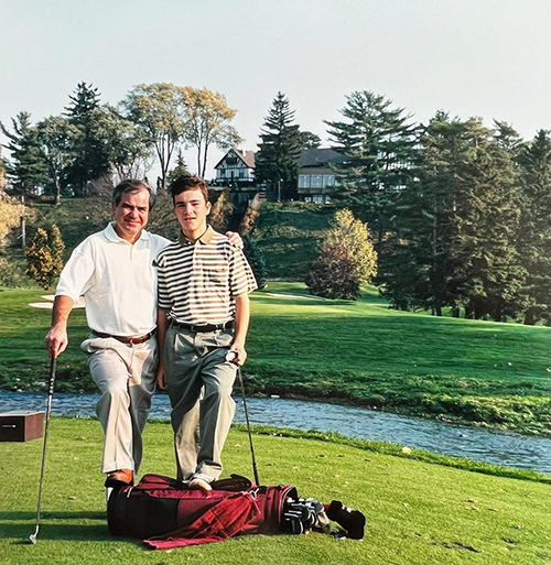 Golf equipment is held by a father and son as trees are in the background.